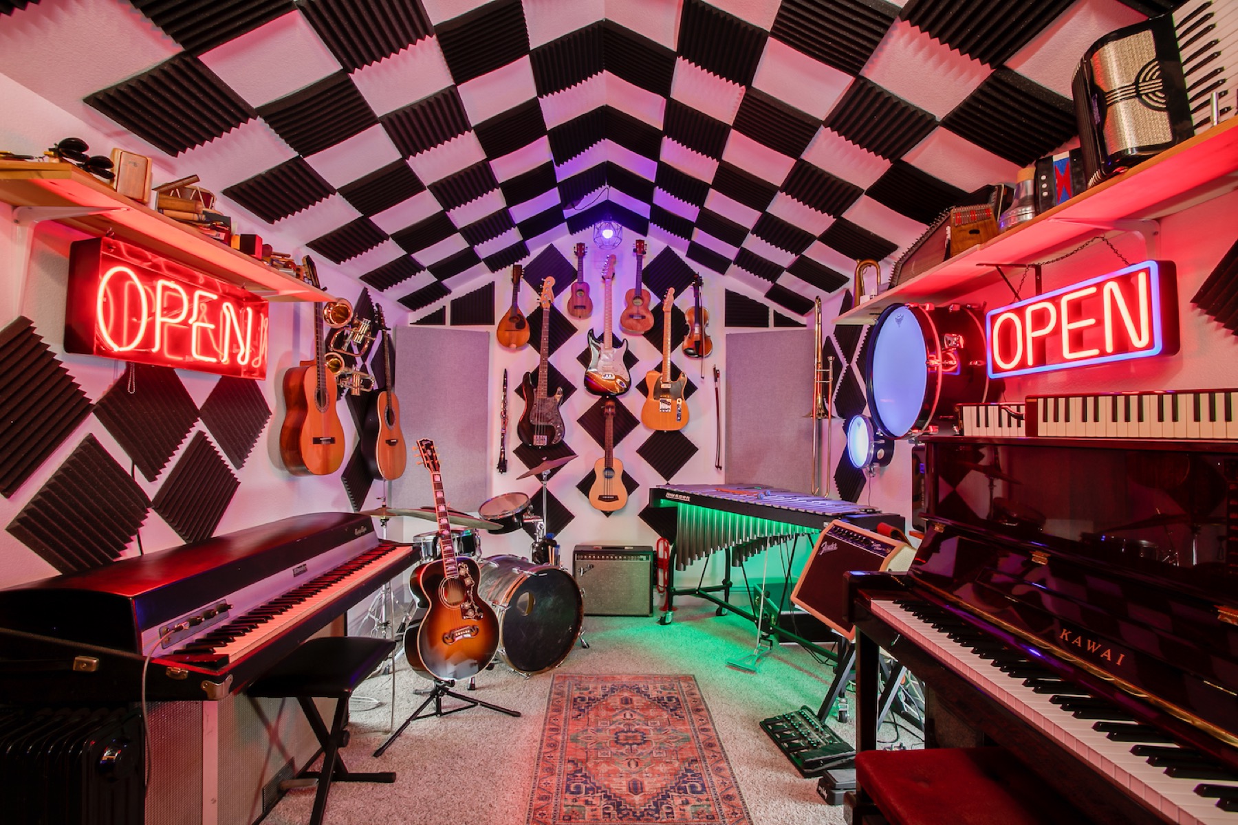My home studio — a checkerboard-foam room lined with guitars, ukuleles, a vibraphone and two pianos, lit in neon red and green.